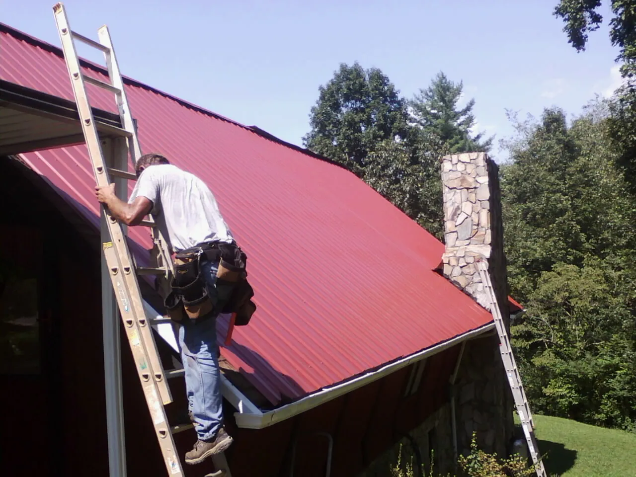 Roofer on a ladder beside a red metal roof and stone chimney — Soffit & Fascia Repair by Roofing Brothers in Moss